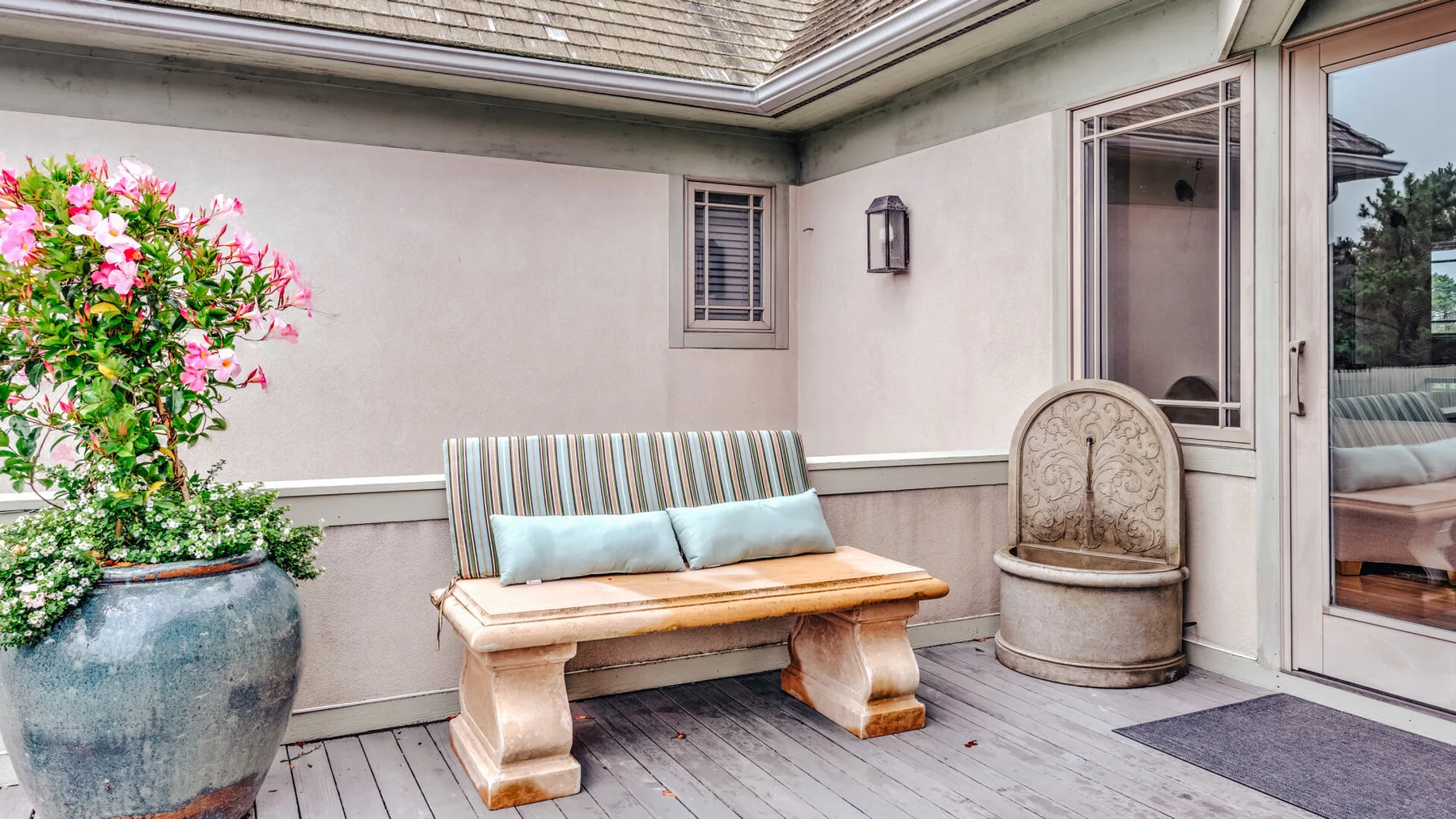 A covered patio has a wooden bench, potted flowers, and a glass door.