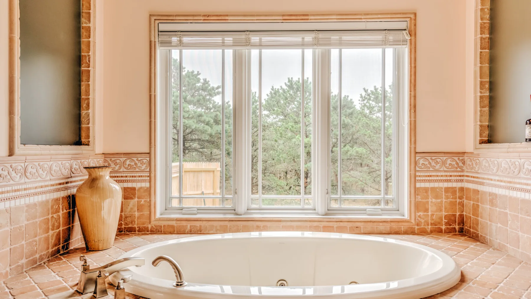 A large bathtub sits beneath a wide window in a tiled bathroom.