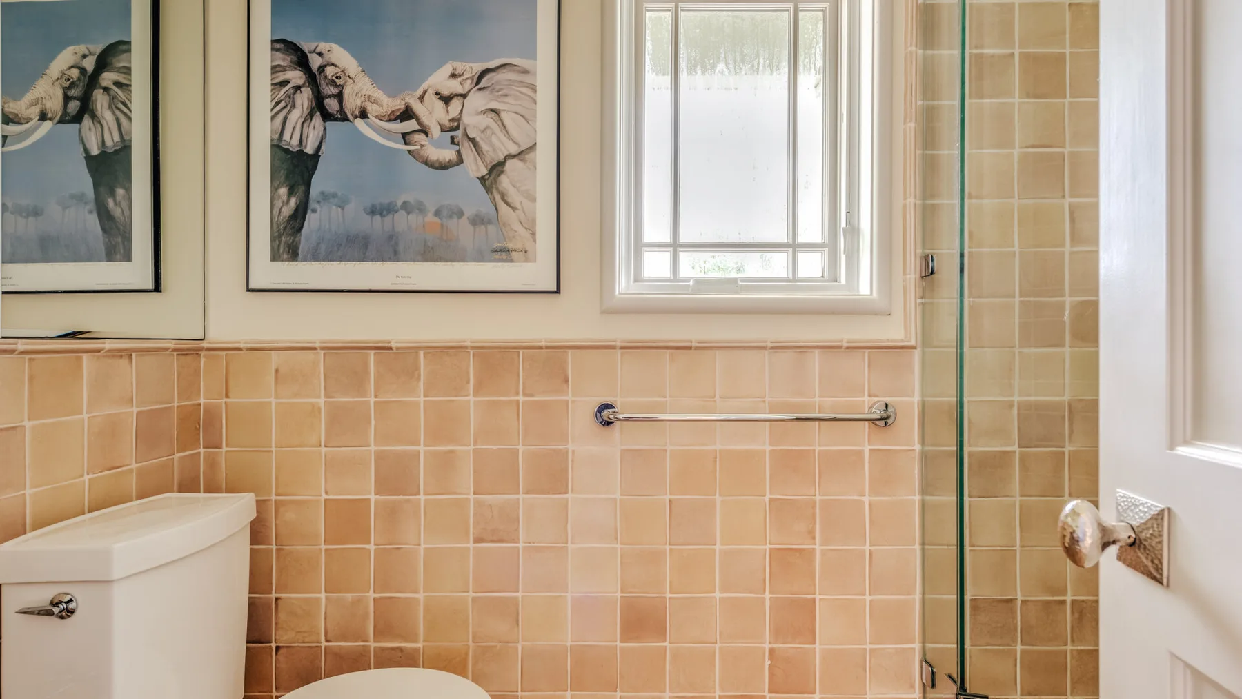 A tiled bathroom with a toilet, shower, and framed elephant artwork.