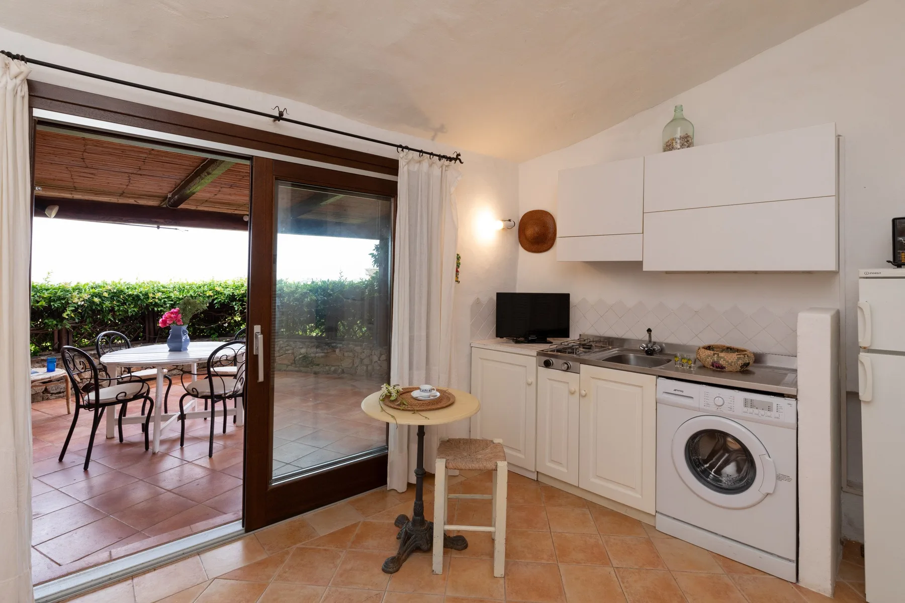 Una cocina compacta con armarios blancos da a un patio embaldosado con mesa y sillas.