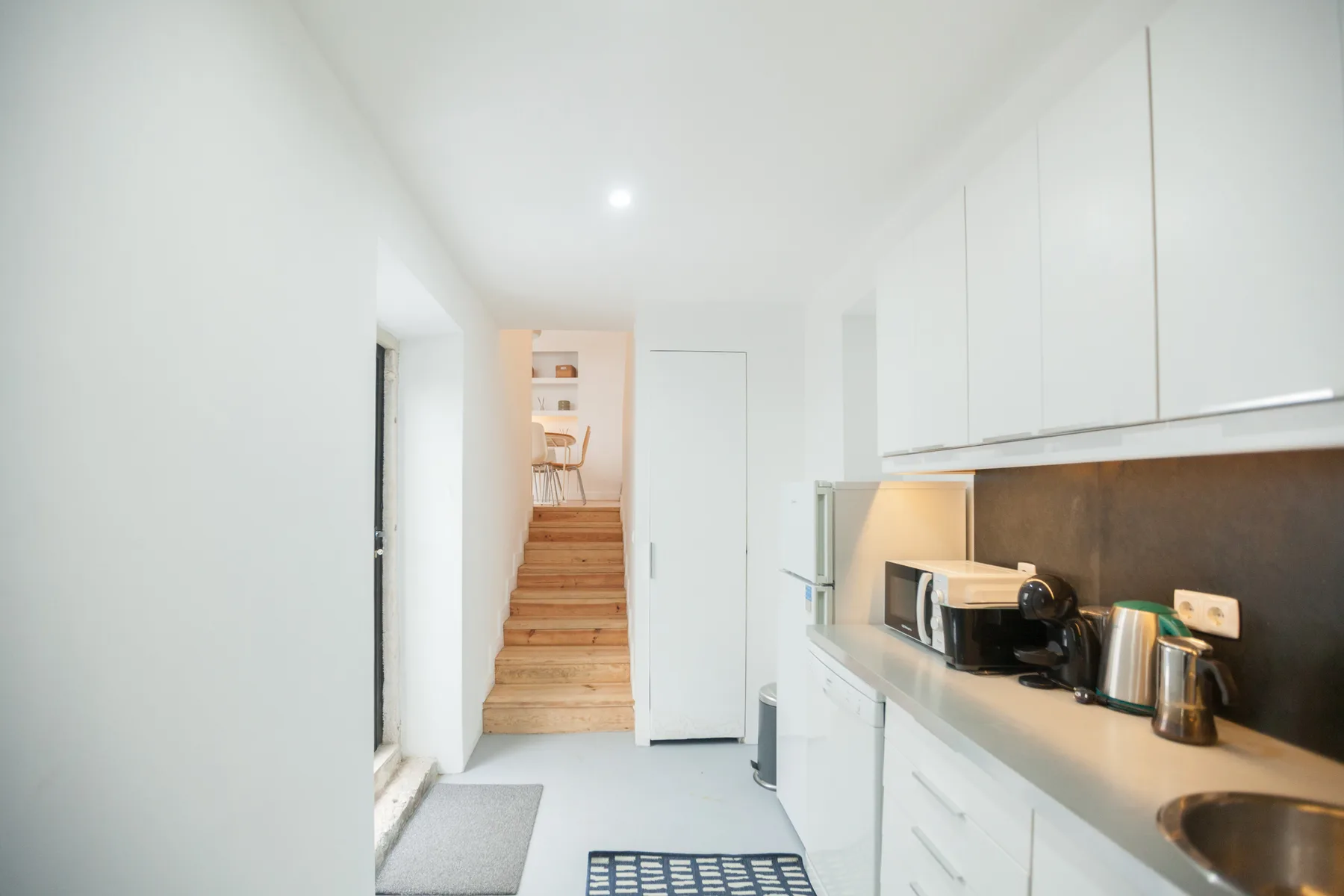 Una cocina blanca y luminosa con una encimera y escaleras de madera al fondo.
