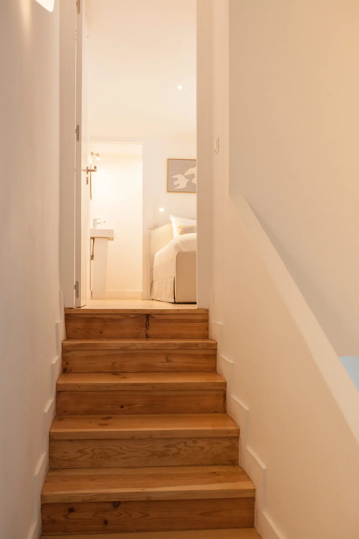 Una escalera de madera conduce a un dormitorio luminoso con una cama y un cuadro.