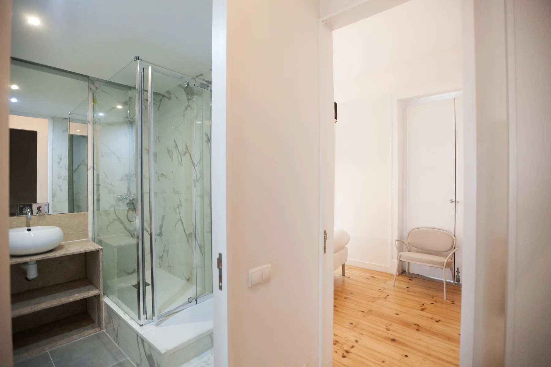 Un baño con ducha de vidrio da a una habitación con suelo de madera y asientos.