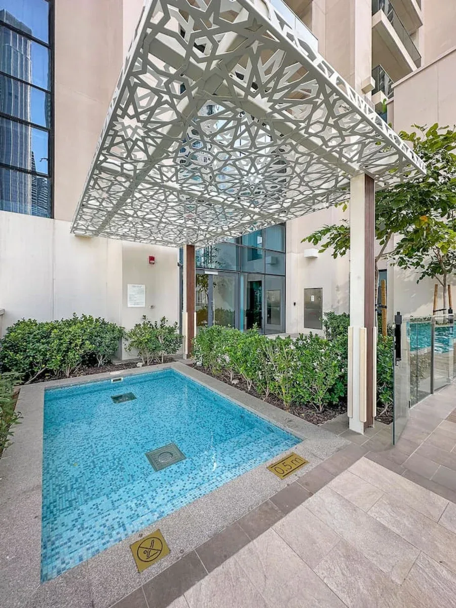 Una pequeña piscina exterior está junto a la entrada de un edificio, bajo un toldo calado.