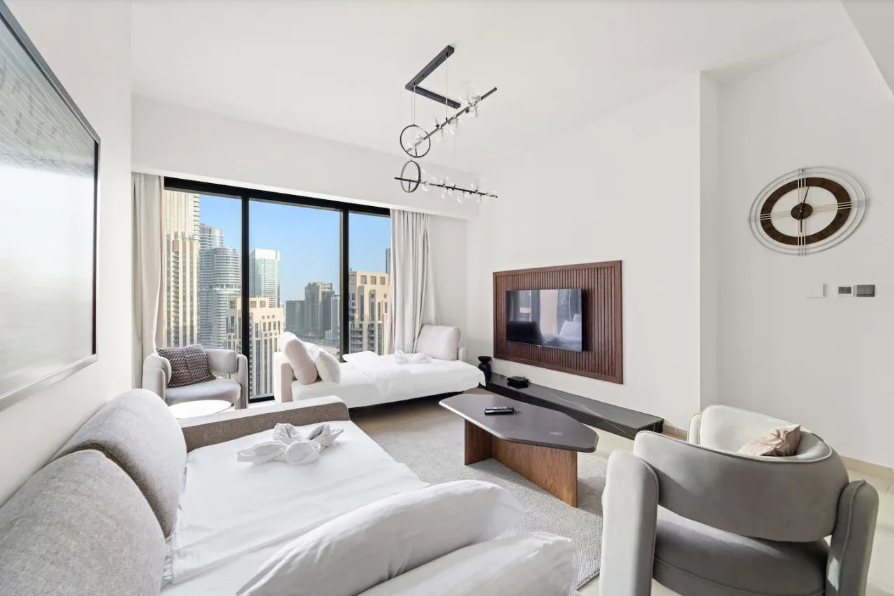 Sala de estar luminosa con asientos blancos, un televisor en la pared y grandes ventanas con vistas a la ciudad.