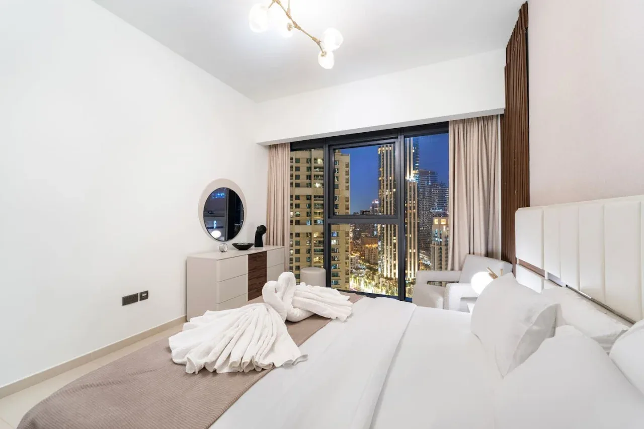 Un dormitorio luminoso con cama, tocador y vistas de la ciudad por grandes ventanales.