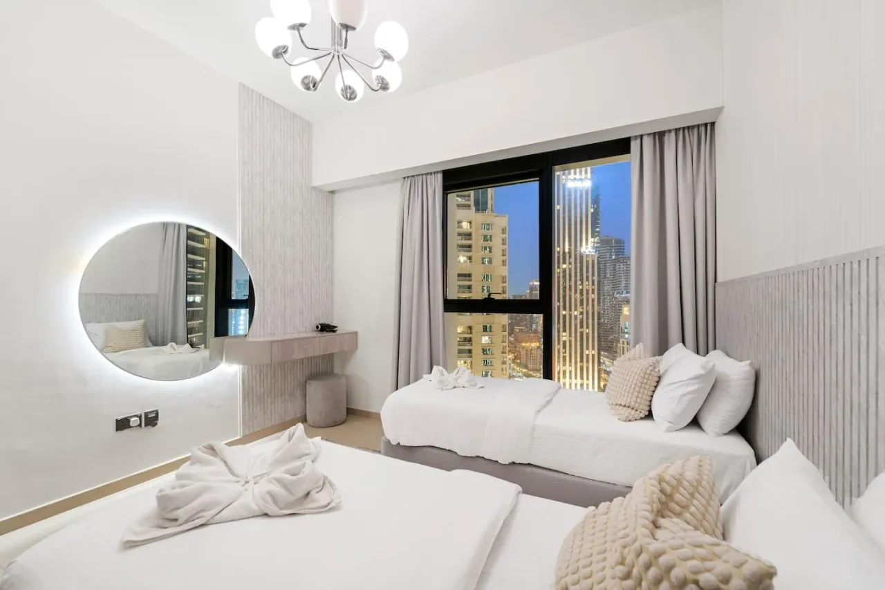 Un dormitorio luminoso con dos camas, un espejo redondo y vistas de la ciudad por la ventana.