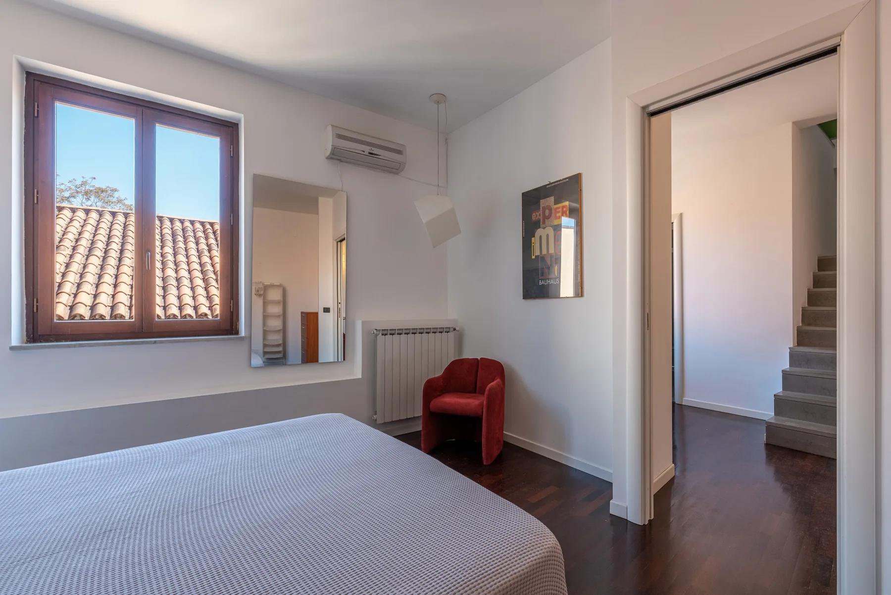 Un dormitorio con una cama, un sillón rojo y una ventana con vista a tejados de tejas.