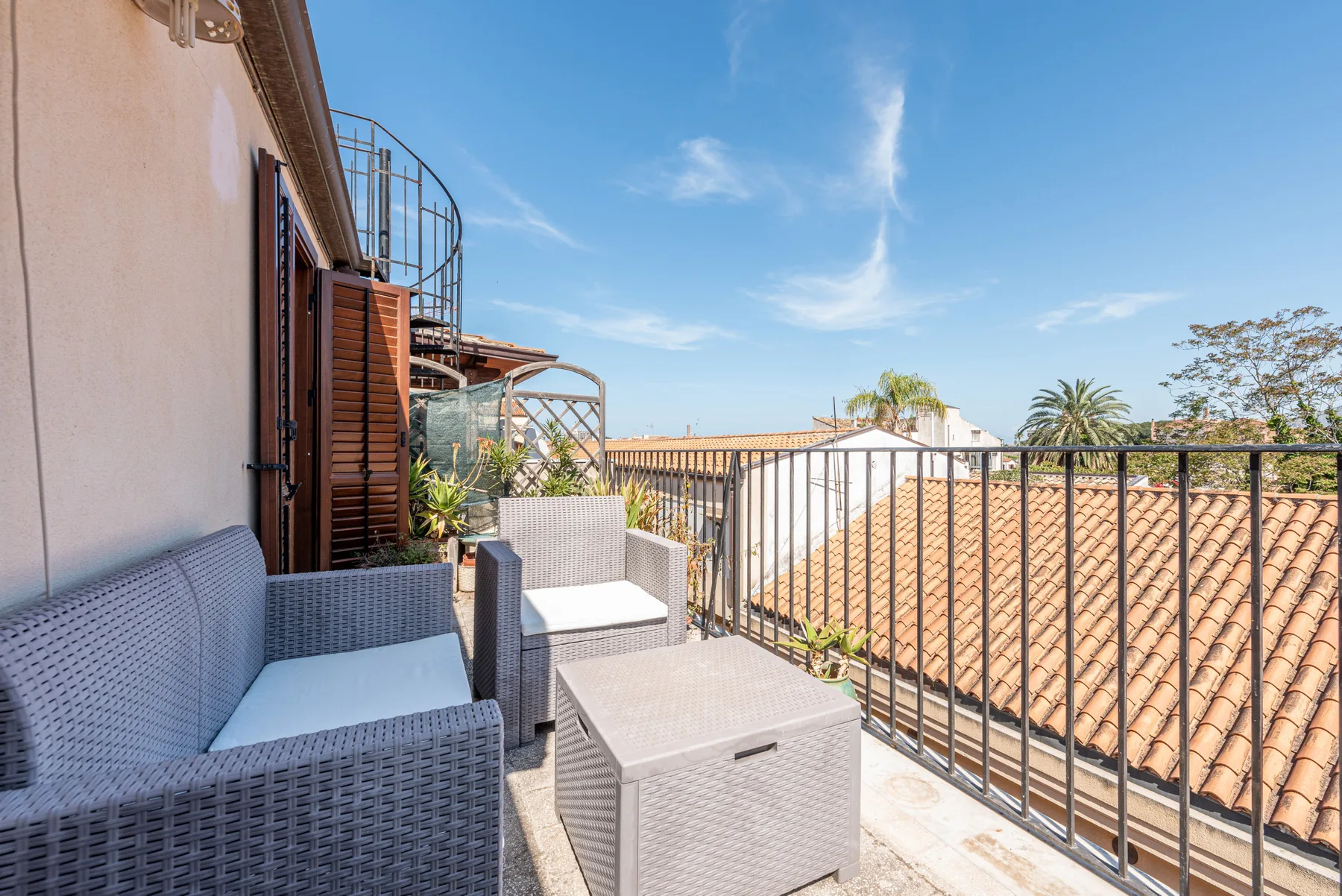 Una terraza exterior soleada con asientos tejidos y una barandilla frente a tejados cercanos.