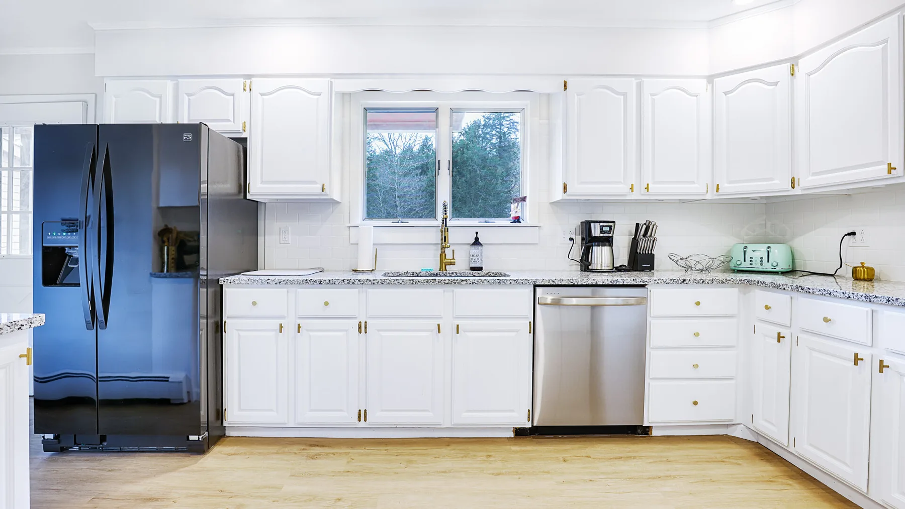 A bright white kitchen with a refrigerator, cabinets, and a window above the sink.