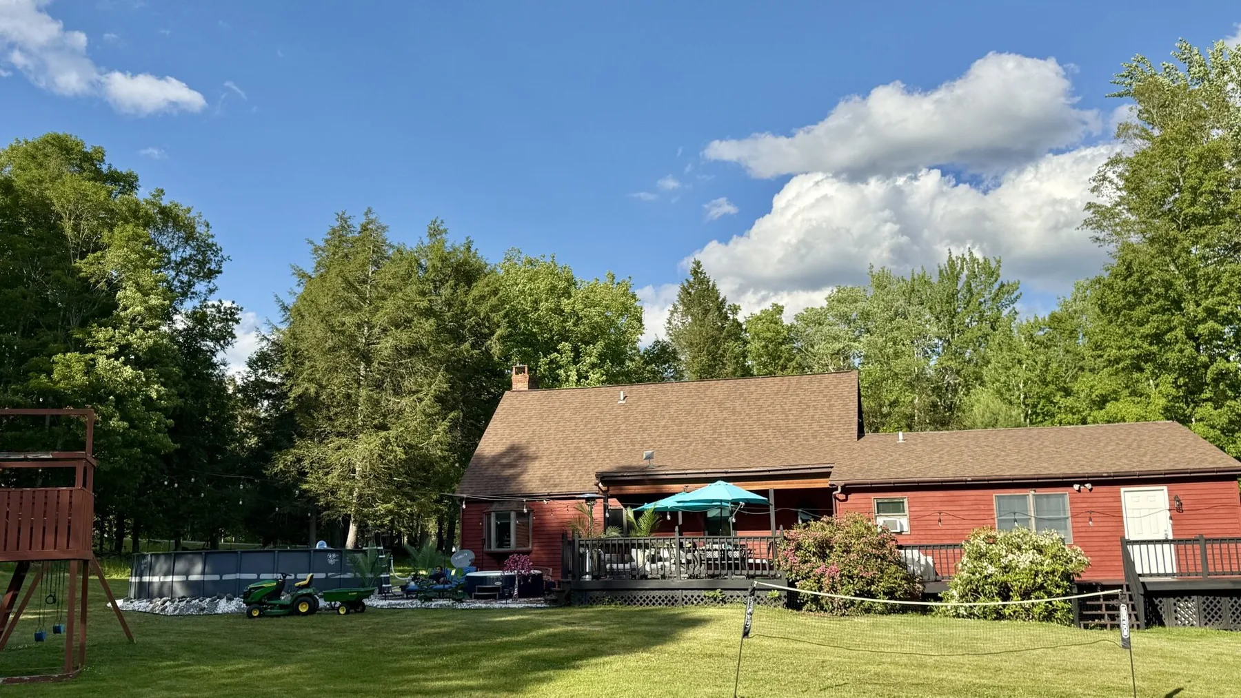 A red house with a broad lawn, patio seating, and trees beneath a blue sky.