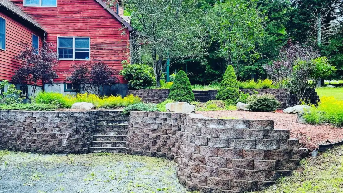A red-sided house stands above a tiered stone retaining wall and landscaped garden.