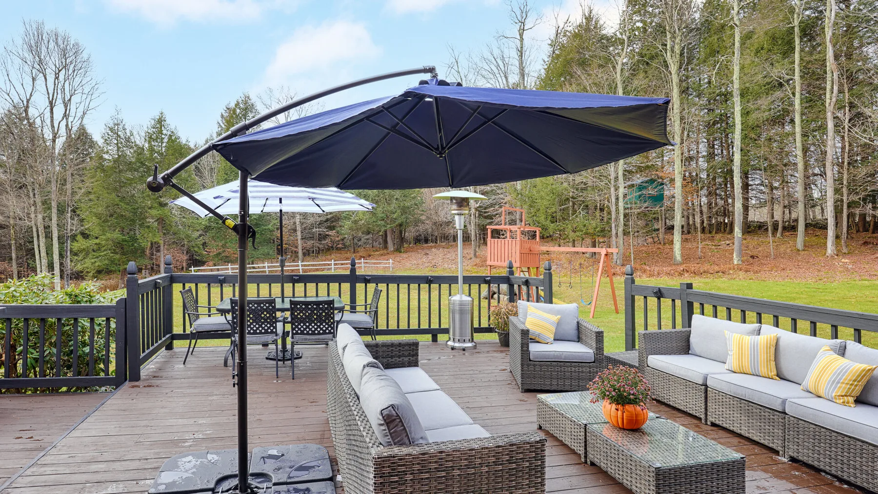 An outdoor deck has seating beneath a large umbrella, overlooking a yard and trees.