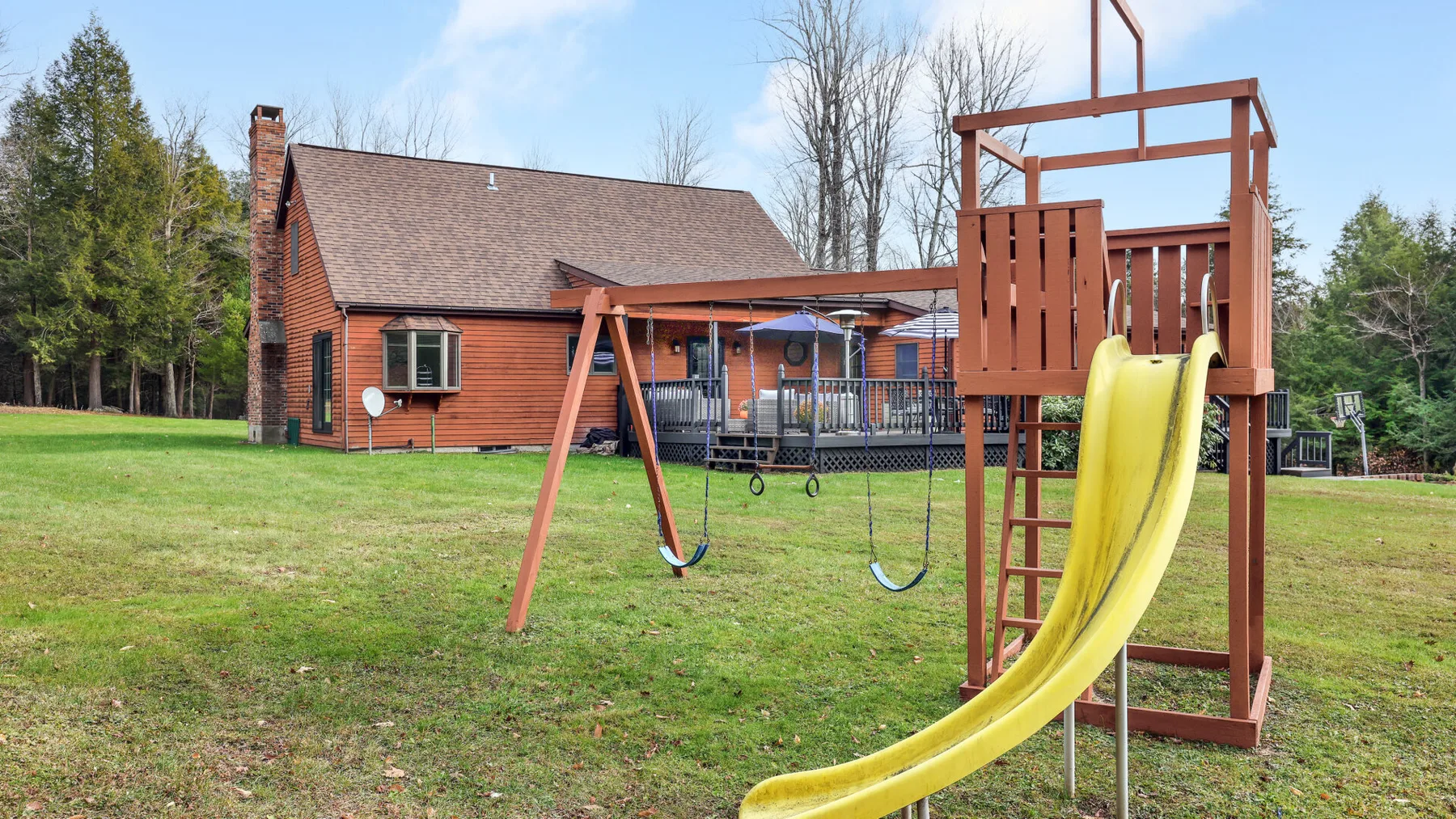 A grassy backyard with a wooden playset, swings, and a yellow slide.