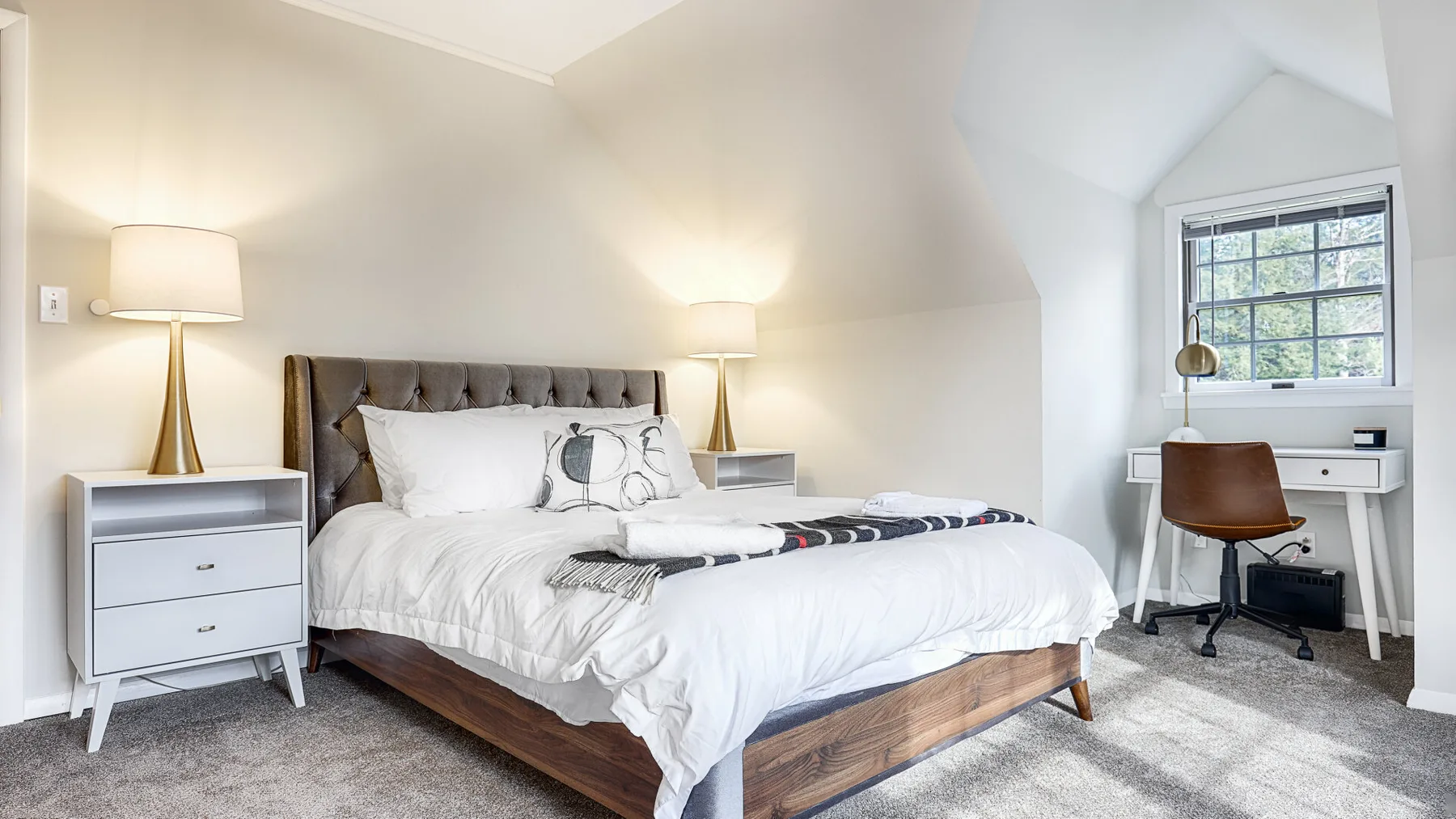 A bright bedroom with a bed, bedside tables, and a desk beneath a window.