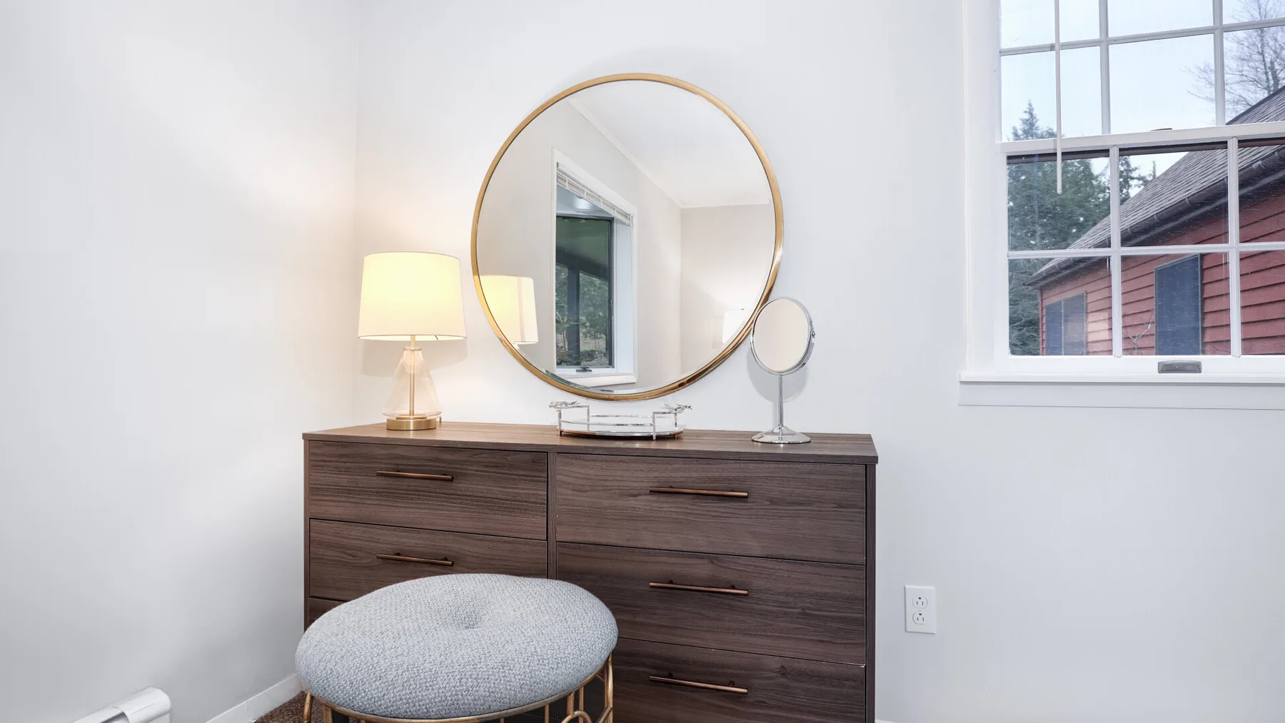 A bedroom dresser stands beneath a round mirror beside a large window.