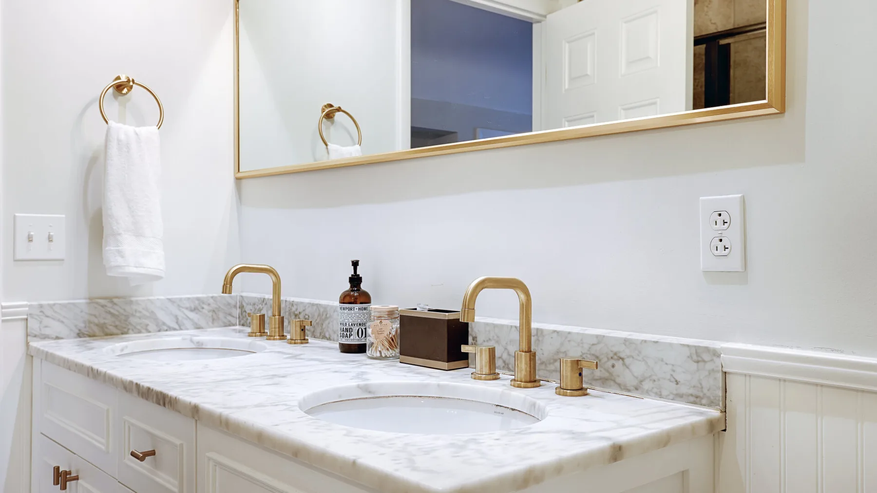 A bright bathroom vanity has a marble-patterned counter, two sinks, and brass faucets.