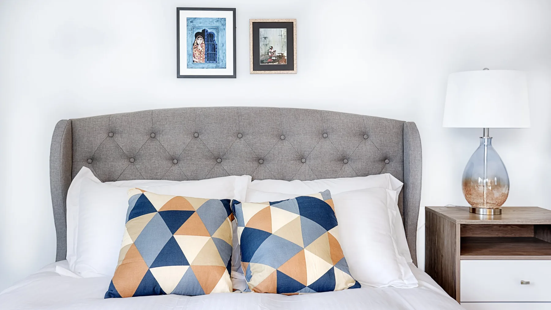 A bedroom with a gray upholstered bed, patterned pillows, and a bedside lamp.