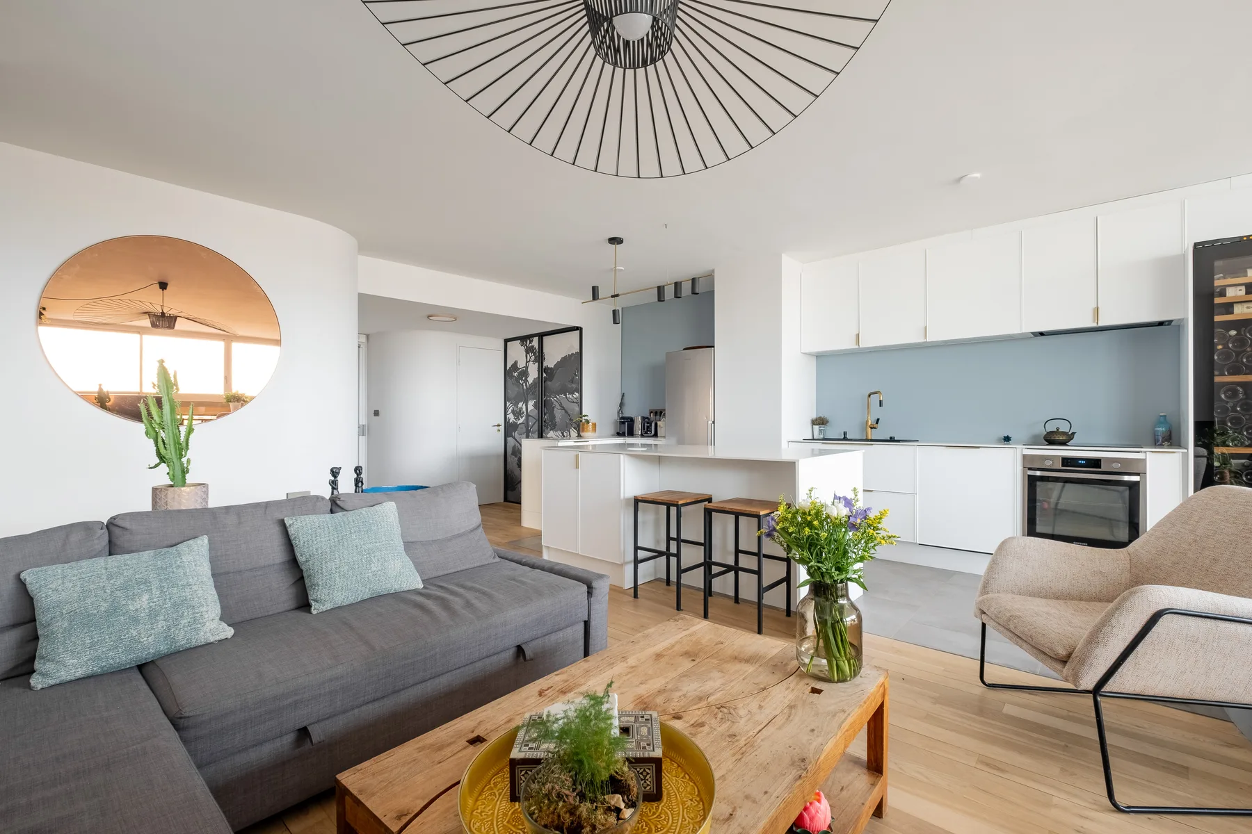 Una sala luminosa con sofá gris, mesa de centro de madera y cocina abierta.