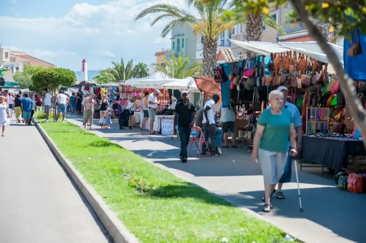 An outdoor market lines a sunny pedestrian walkway with clothing stalls and shoppers.
