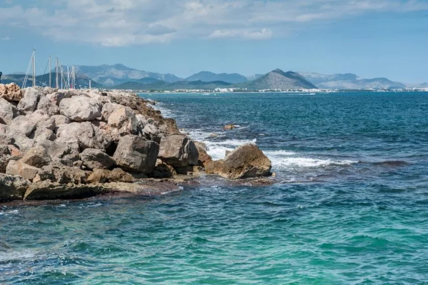 A rocky coastline borders clear blue water, with distant mountains across the bay.