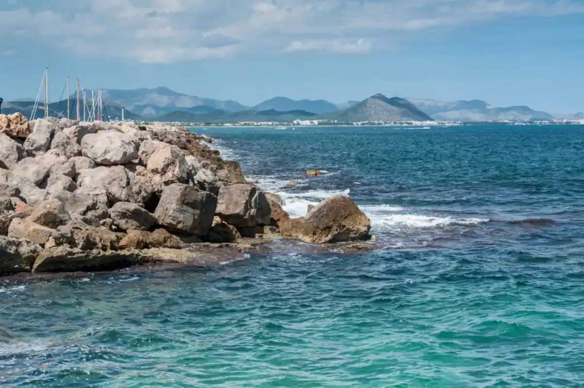 Rocky coastline beside blue water, with distant mountains under a cloudy sky.