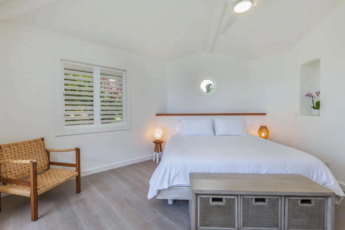 A bright bedroom with a bed, woven seating, and a window with shutters.