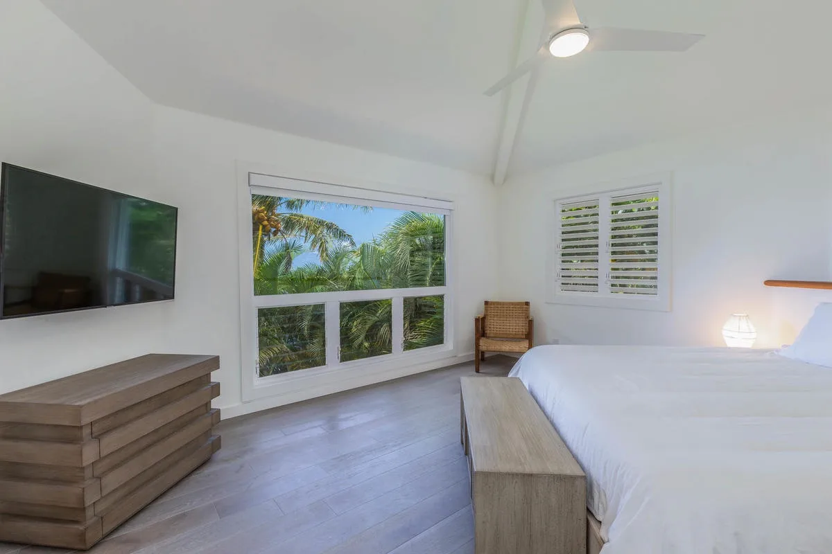A bright bedroom with a large bed, a wall-mounted television, and windows overlooking greenery.
