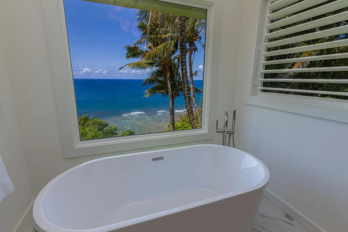 A bathtub sits beside a window overlooking palm trees and the ocean.