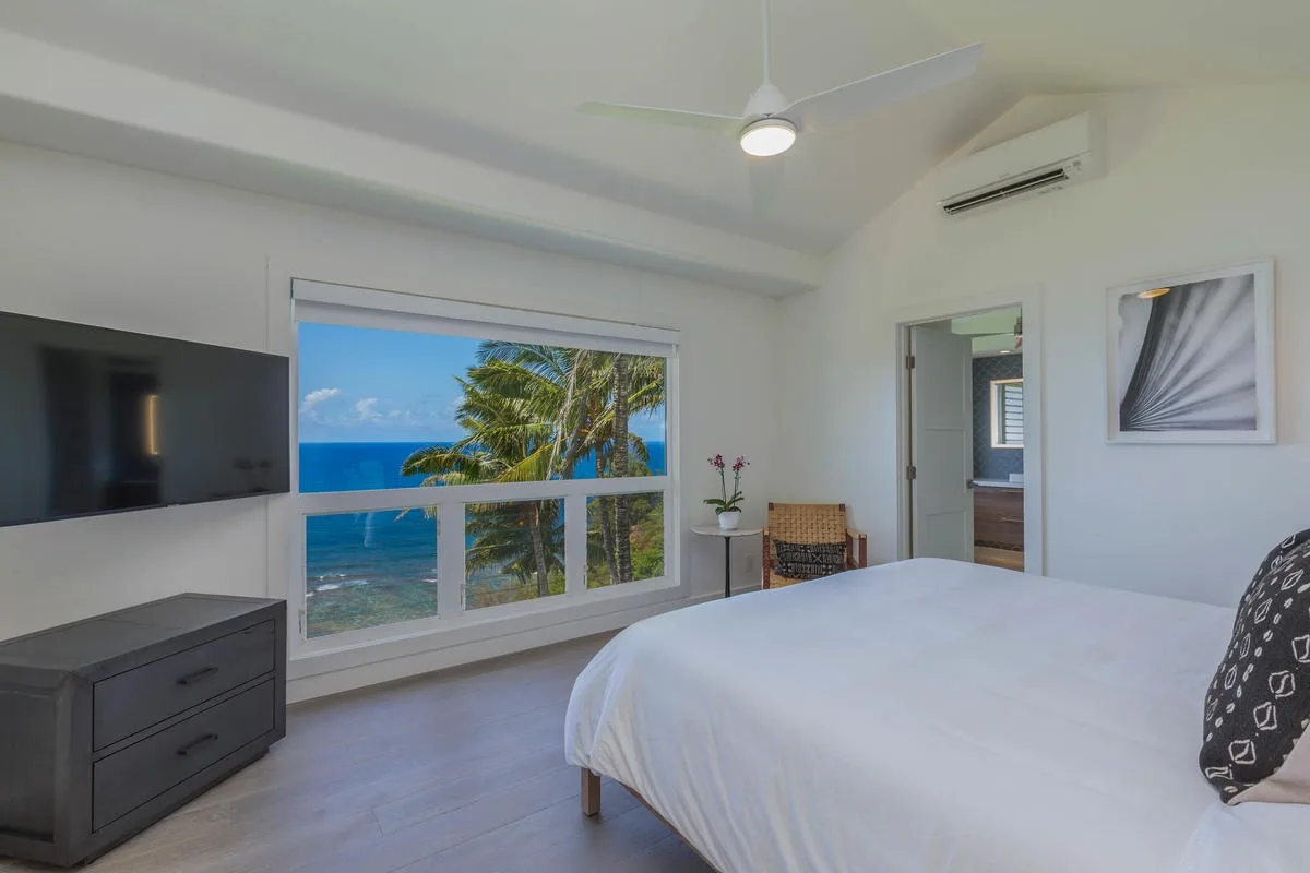 A bright bedroom with a large bed and a wide window overlooking the ocean.