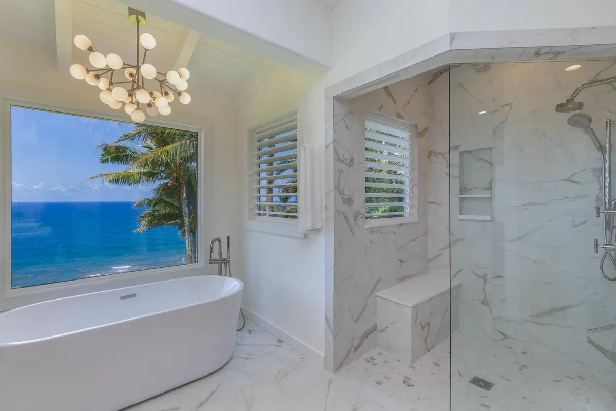 A bright bathroom has a freestanding tub beside a large window overlooking the ocean.