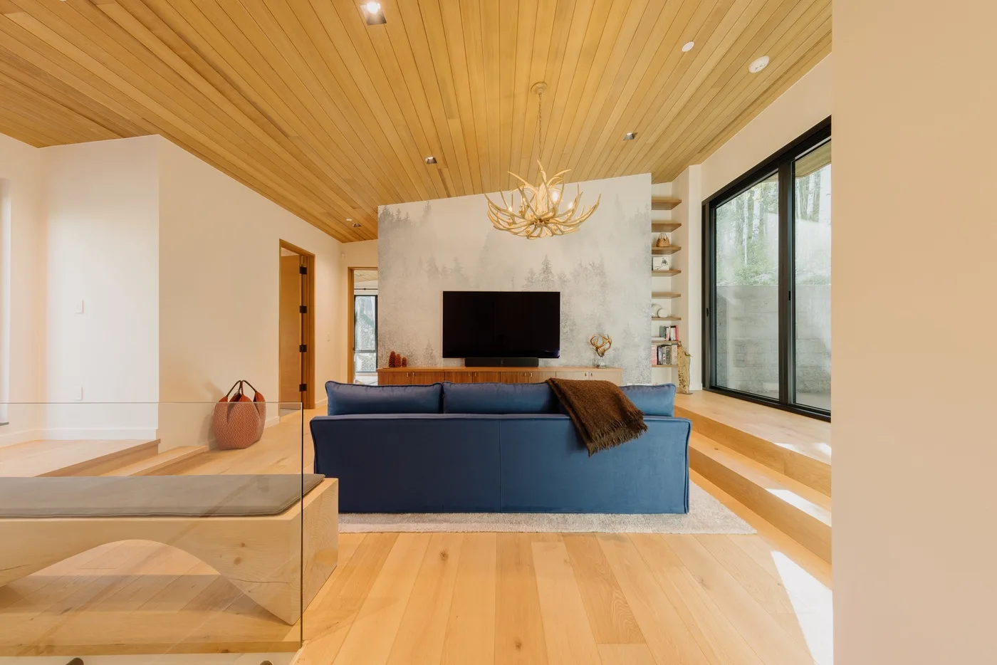 Una sala de estar con asientos azules, un televisor y ventanales.