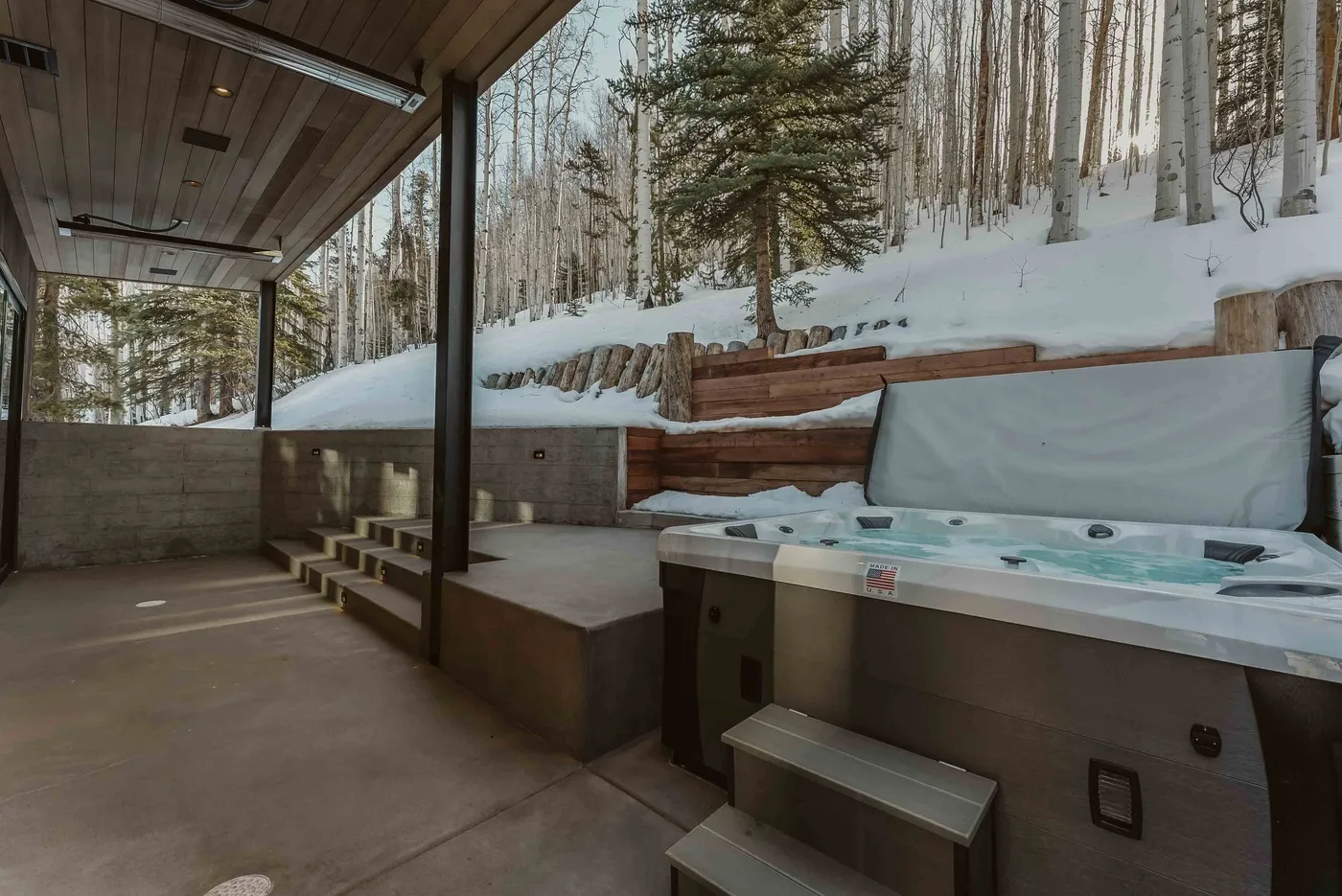 Una bañera de hidromasaje al aire libre está bajo un patio cubierto junto a una ladera boscosa nevada.