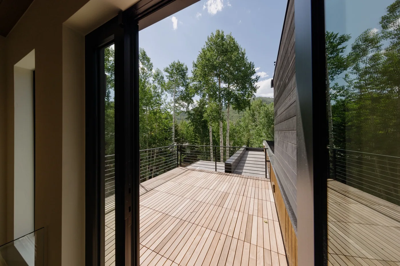Una terraza de madera se extiende tras unas puertas de vidrio, frente a árboles frondosos.