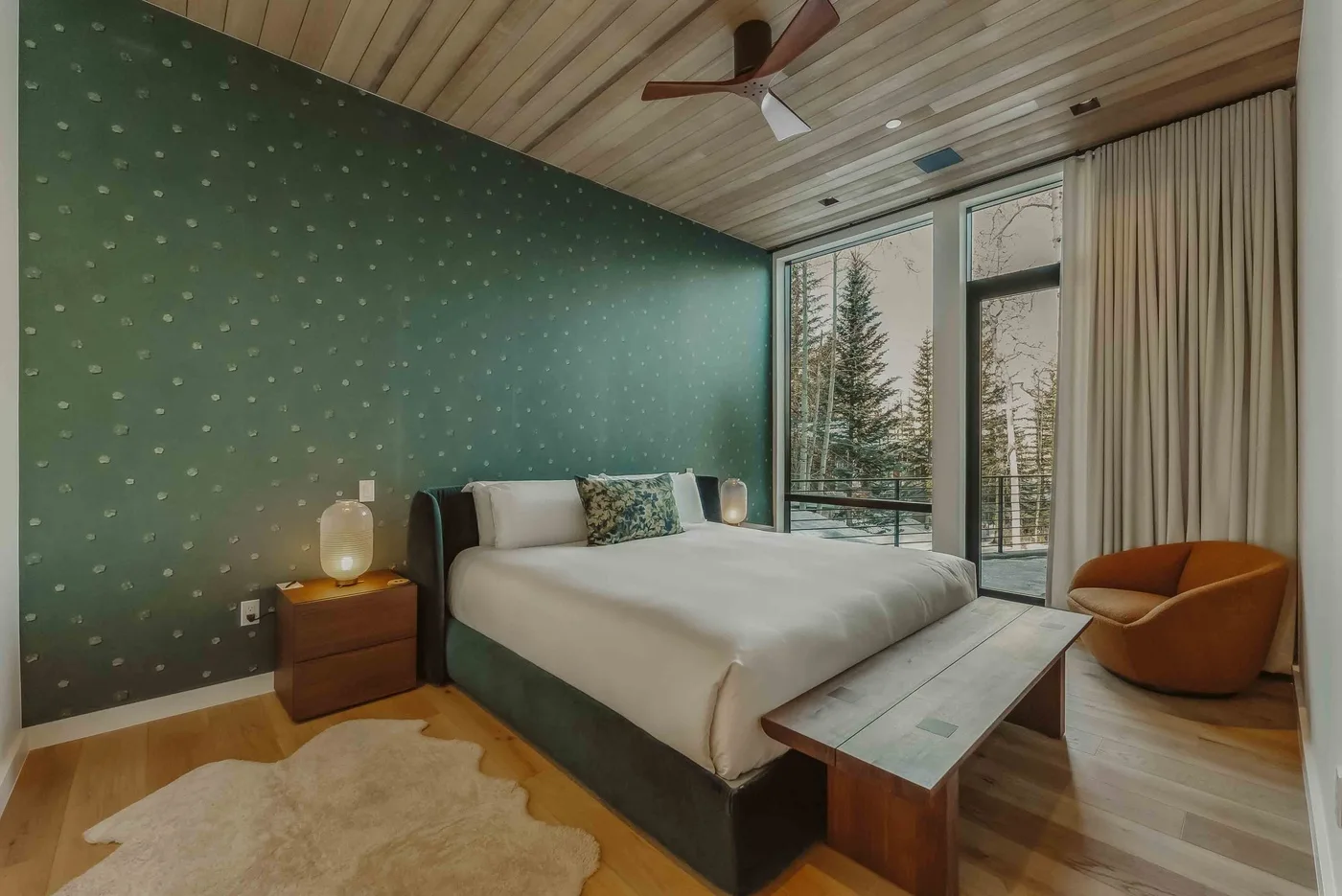 Dormitorio con una cama, un asiento naranja y ventanas altas con vistas a los árboles.