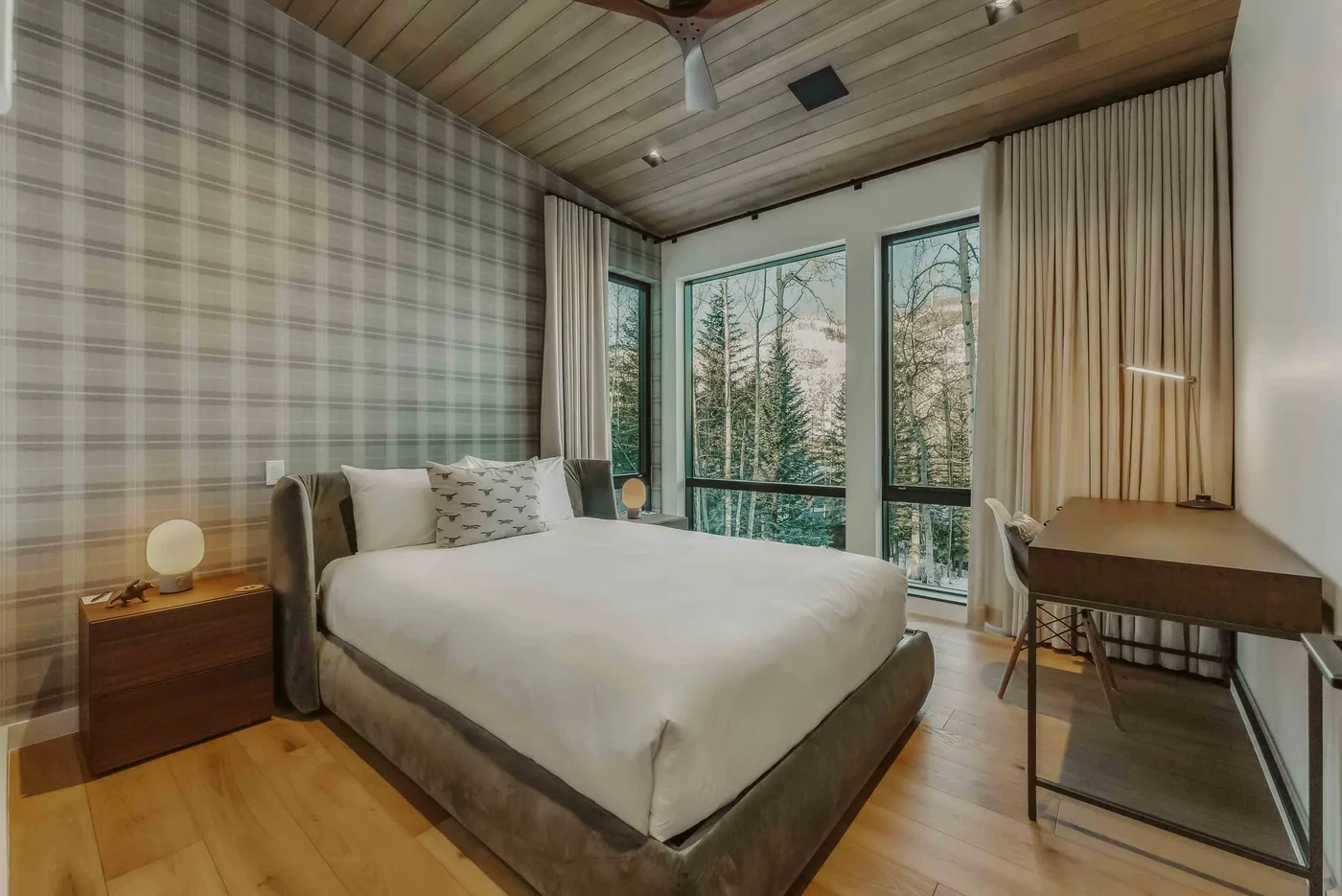 Un dormitorio con una cama, una mesita de noche y grandes ventanas con vista a los árboles.