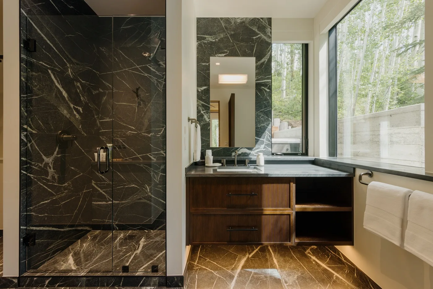 Un baño moderno tiene una ducha de piedra oscura, un tocador de madera y ventanas grandes.