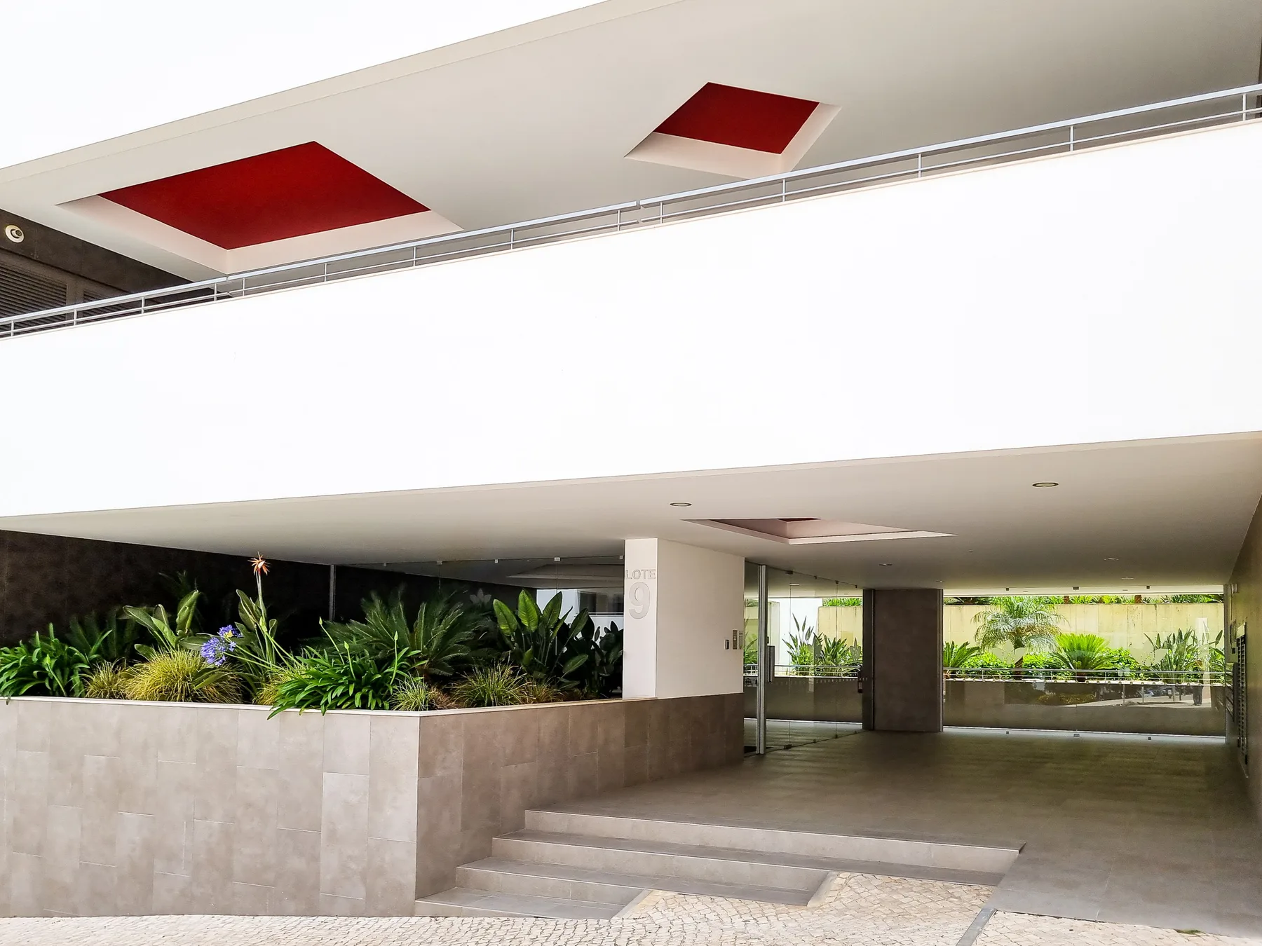 A covered building entrance opens onto a broad paved walkway beside planted beds.