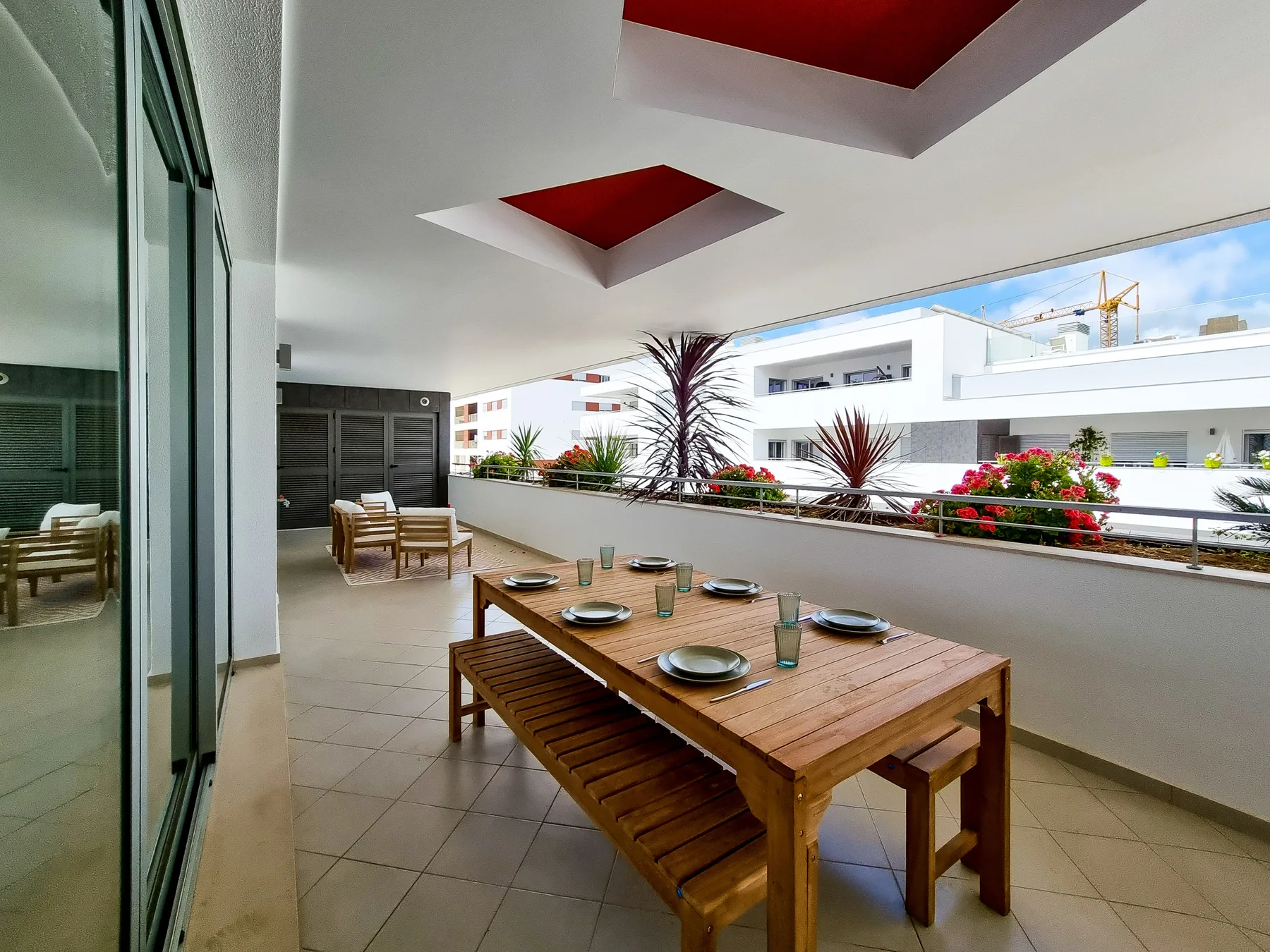 A covered terrace with a wooden dining table overlooks nearby buildings and greenery.