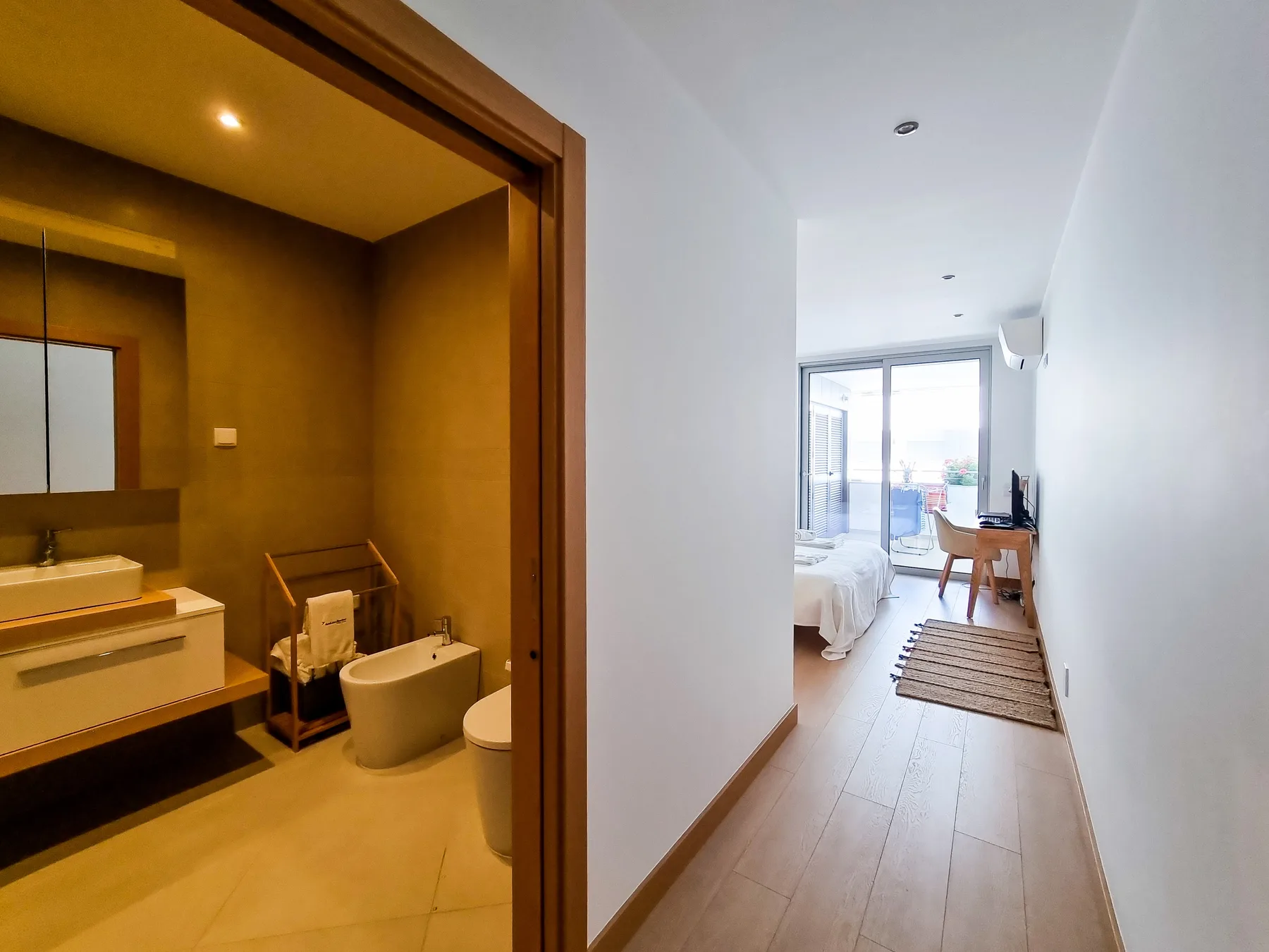 A bathroom opens onto a bright bedroom with a desk and large windows.
