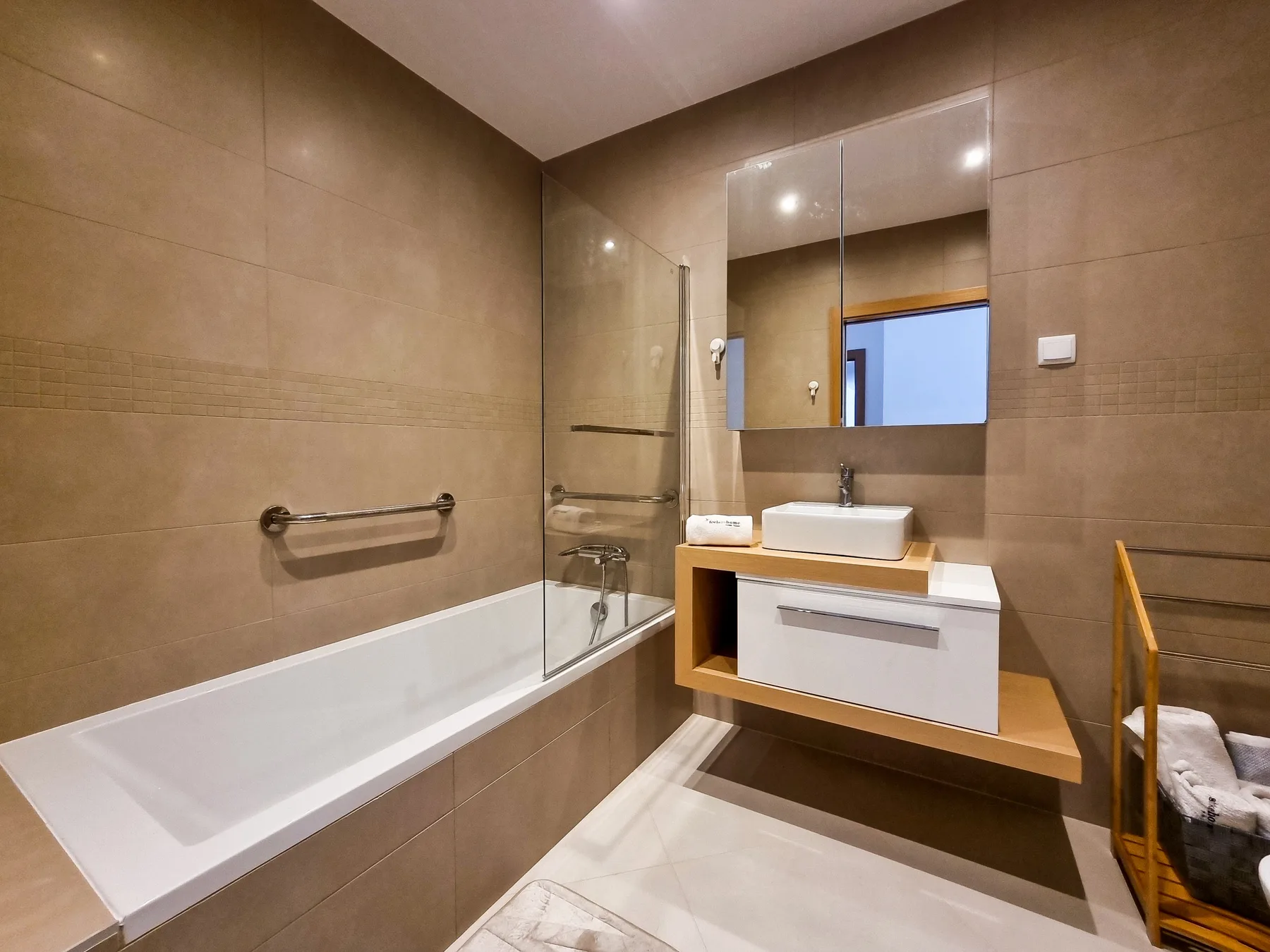 A tiled bathroom with a bathtub, glass shower screen, and wall-mounted sink.