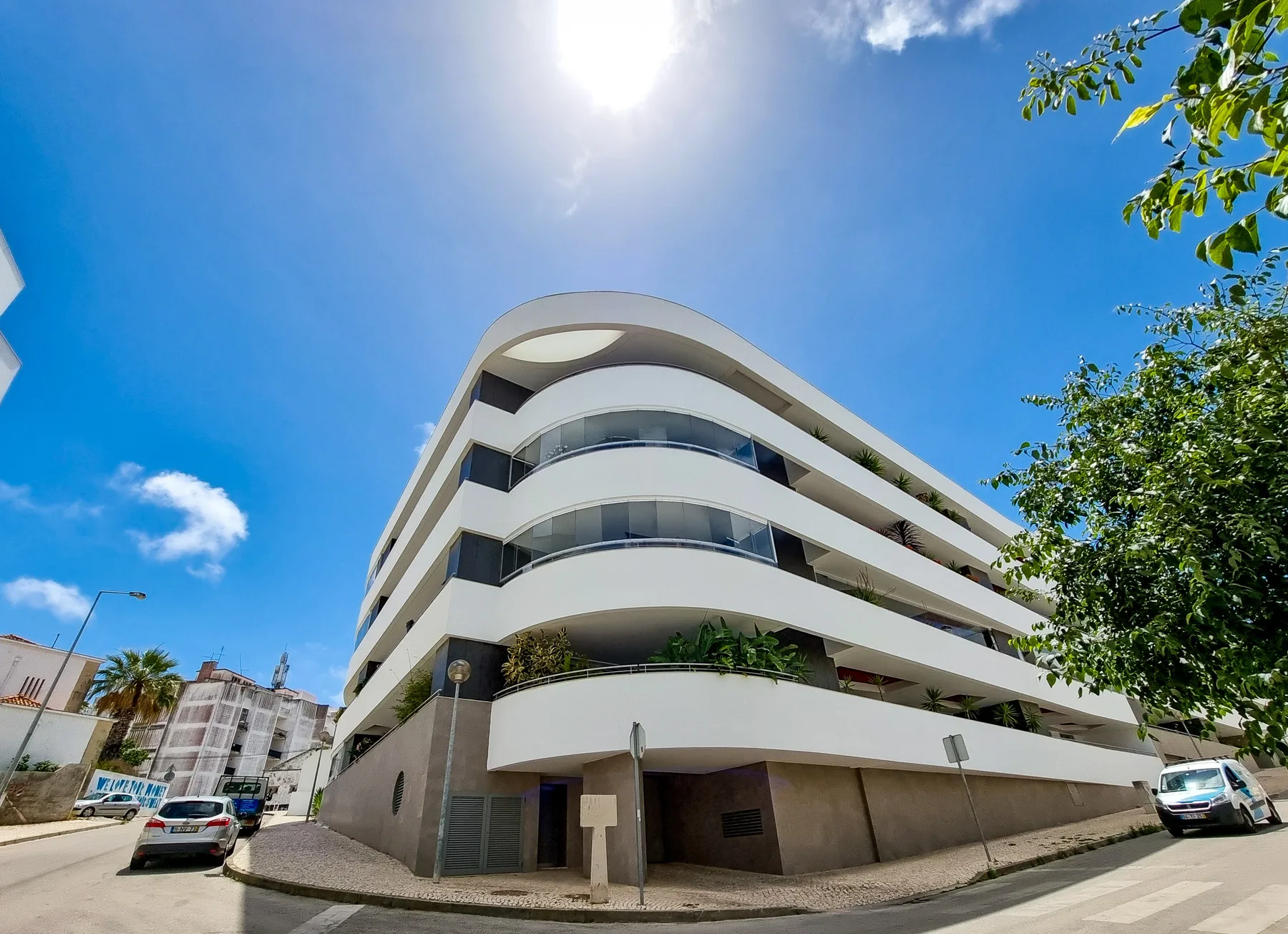 A curved modern building with stacked balconies stands at a sunny street corner.