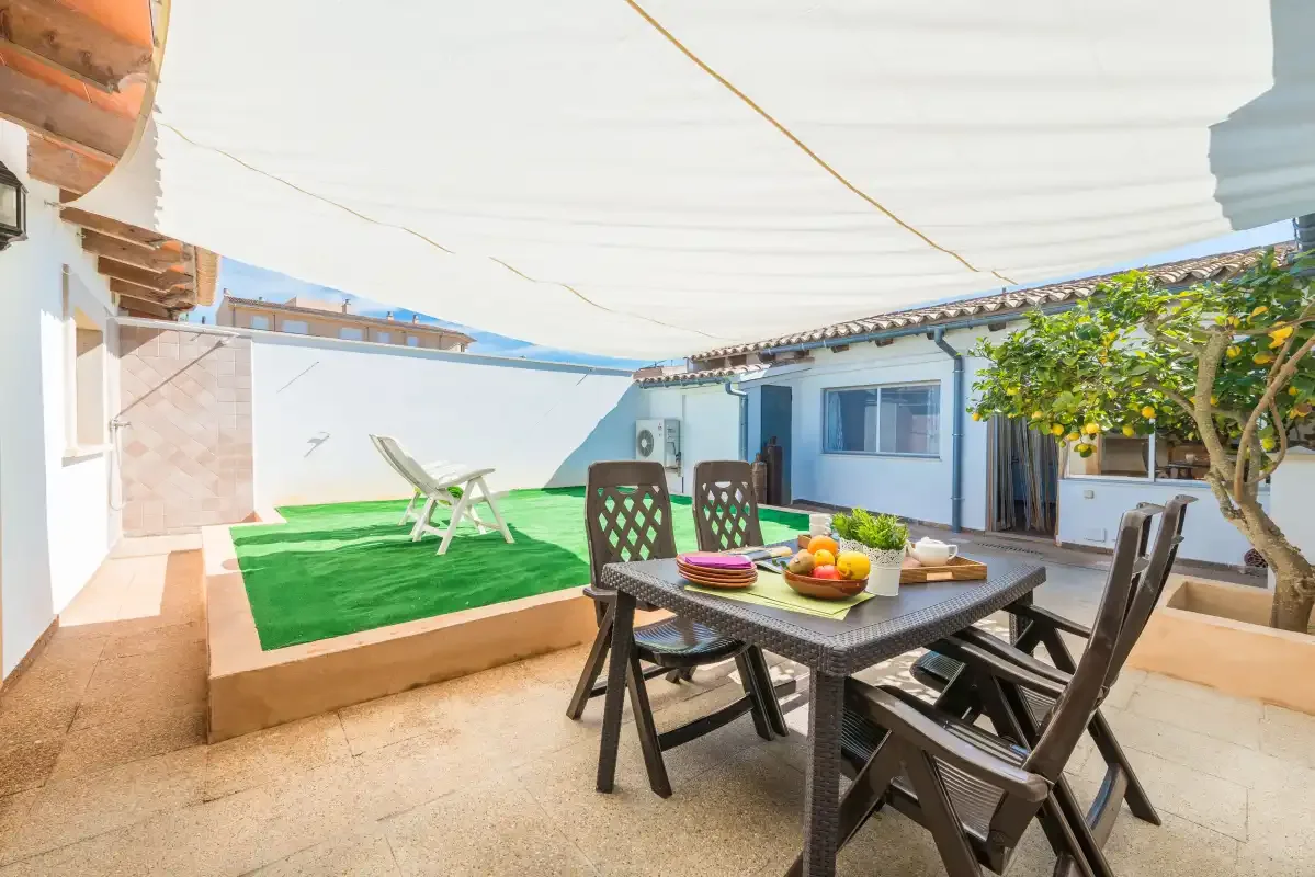 A shaded outdoor dining area with a table and chairs beside a small green lawn.