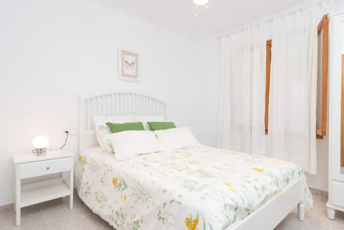 A bright bedroom with a bed dressed in floral bedding and white curtains.