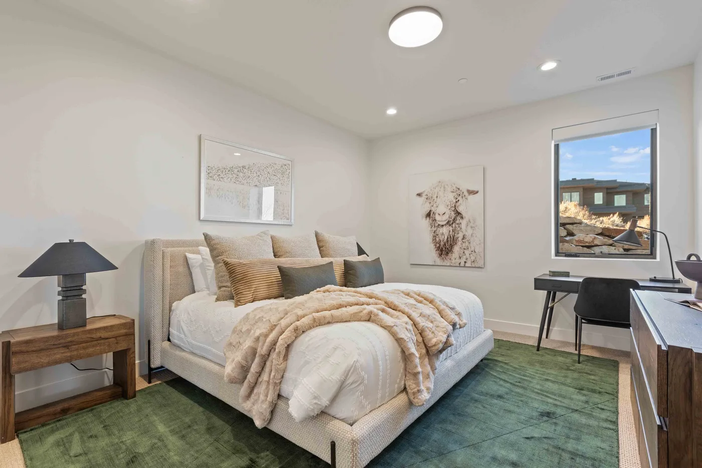 A bedroom with a bed, bedside table, and a window overlooking a neighboring building.
