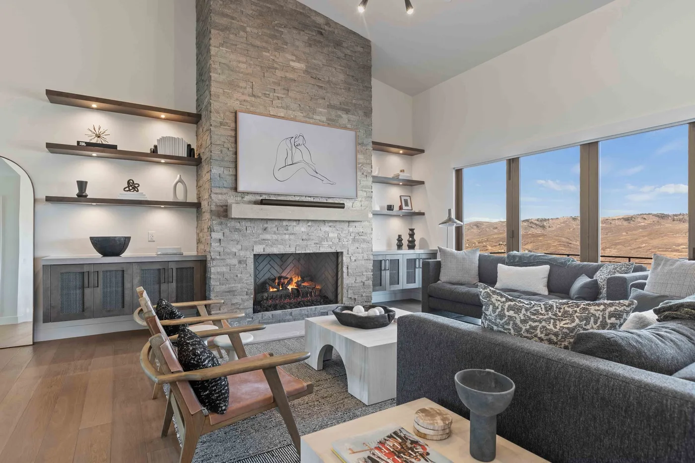 A living room with a stone fireplace, seating, and large windows overlooking a dry landscape.