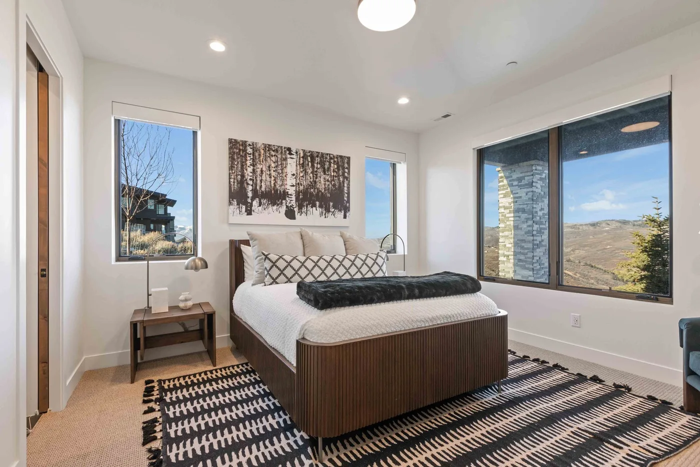 A bright bedroom with a bed, patterned rugs, and large windows overlooking the landscape.