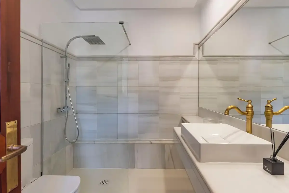 A bathroom with a glass shower and a sink topped by gold-colored faucets.