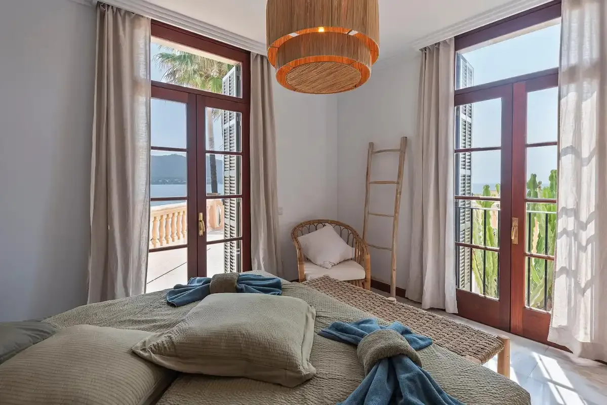 A bright bedroom with a bed, a small seating area, and tall windows.