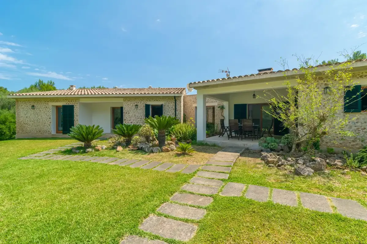 Una casa de piedra da a un jardín con césped y un sendero hacia el patio cubierto.